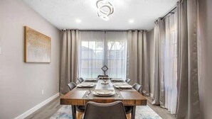 A dining room with a long wooden table set for six, surrounded by chairs, with a modern chandelier above and a large window with curtains. - Luxury Home 4 Rooms ~ Pool Table & Poker~ King Bed (Houston)
