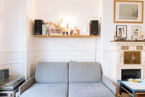 A room with a grey sofa, a wooden shelf with books, a fireplace, and a framed picture on the wall. - Central, Spacious Family Apartment with Balcony (Paris)