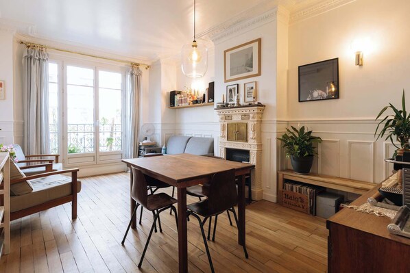 A room with a wooden dining table, chairs, a fireplace, a bookshelf, and a window with curtains.