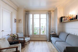 Ein Zimmer mit Holzboden, einem grauen Sofa, einem Holzstuhl, einem kleinen Tisch mit Blumenvase und einem BĂŒcherregal mit BĂŒchern und Dekoration.