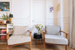 Two armchairs with cushions, a small round table with a vase of flowers, and a bookshelf with books. - Central, Spacious Family Apartment with Balcony (Paris)