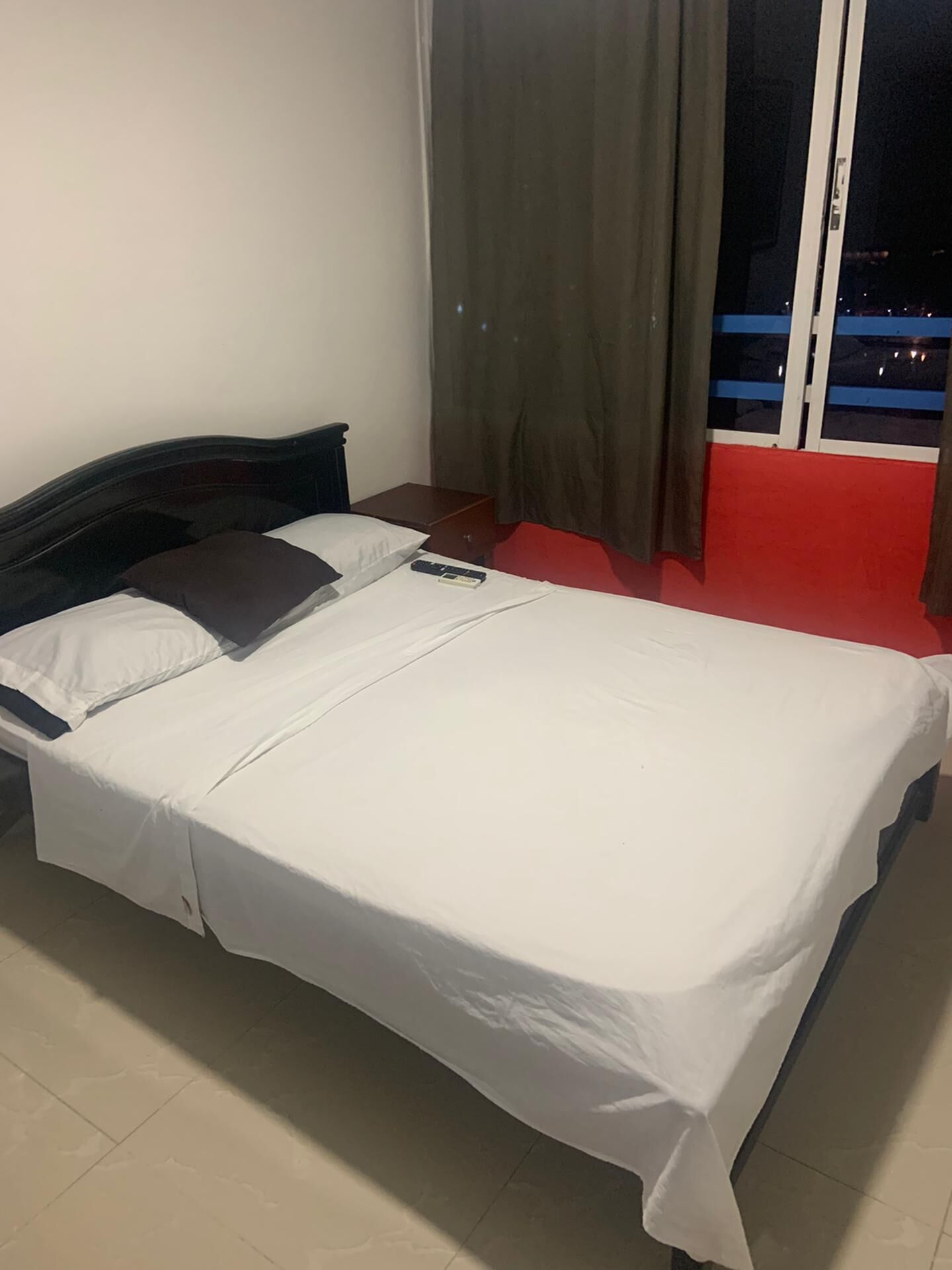 Un lit simple avec une literie blanche, une tête de lit en bois, une table de chevet et une chaise rouge.