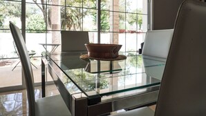 Uma mesa com tampo de vidro, um vaso de terracota, uma cadeira branca e vista para um pátio exterior através de grandes janelas.
