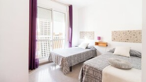 Une chambre avec deux lits, une fenêtre avec des rideaux violets et une étagère fixée au mur, sur laquelle se trouve une lampe.