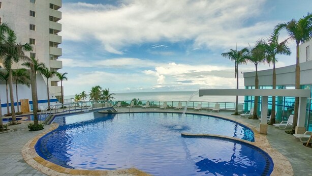 A pool area with a view of the ocean, surrounded by palm trees and lounge chairs. - Apartamento Al-4 Frente al mar Piscina y Wifi (Cartagena)
