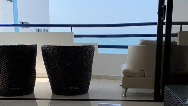 A balcony with a white sofa and two wicker chairs, a glass door leading to a pool, and a view of a building with blue and white exterior. - 3mr8 Duplex Apartment Close To The Sea With Pool (Cartagena)