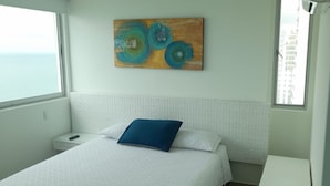 A bedroom with a bed, a nightstand, a painting, and a window with a view of the sea.