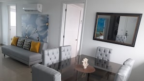 A modern living room with a grey sofa, two grey armchairs, a glass coffee table, and a large mirror reflecting a beach scene.