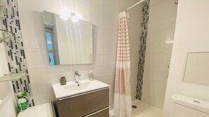 Et moderne badeværelse med en hvid vask, spejl og toilet. Der er en bruser med blomstret gardin og bruseafskærmning i glas.