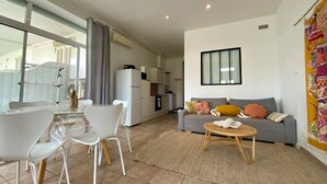 A modern living room with a grey sofa, white chairs, and a small round table. There is a kitchen area in the background with a refrigerator and a window.