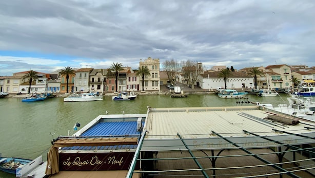 A marina with boats docked, a building with a sign reading "Le Duc Dix - Nouveau", and a cloudy sky.
