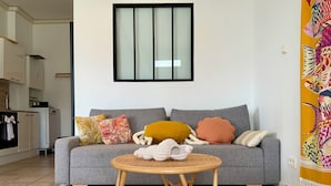 A modern living room with a grey sofa, colorful cushions, a small round wooden table, and a kitchen in the background.