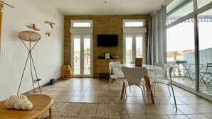 A spacious living area with a dining table, chairs, a TV mounted on the wall, and a large window offering a view of the outdoors.