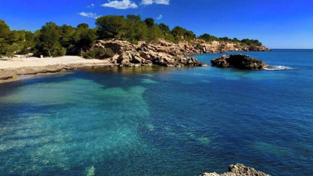 A coastal landscape with clear blue waters, rocky outcrops, and dense greenery.