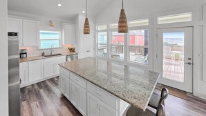 A kitchen with white cabinets, a granite countertop, hanging wicker pendant lights, and a view of a red building through large windows. - Breezy Ocean View/Sleeps 10/Steps To Beach/Central (Bolivar Peninsula)