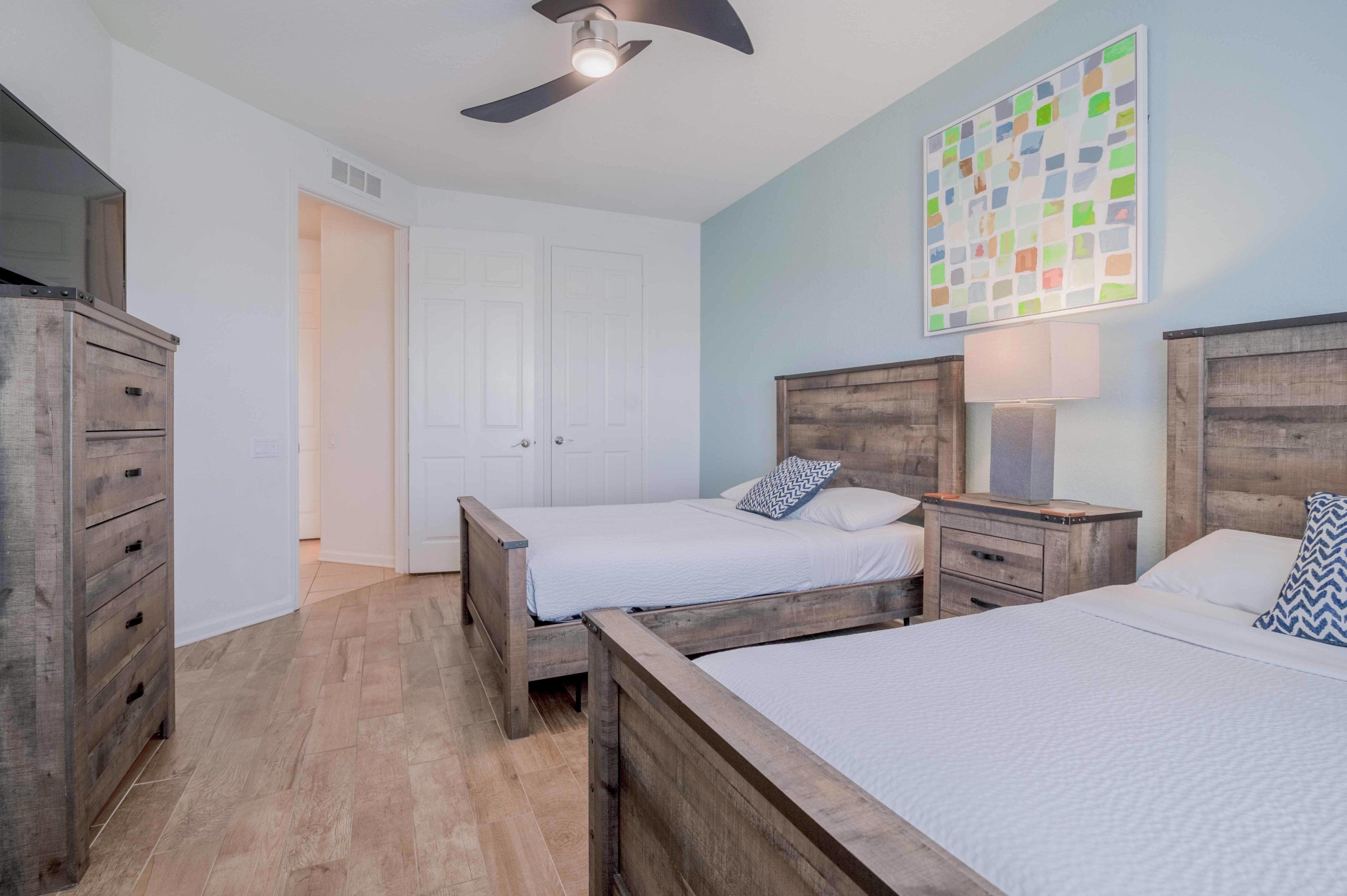 Une chambre à coucher avec deux lits en bois, un ventilateur de plafond, une porte blanche et une peinture abstraite aux couleurs vives accrochée au mur.
