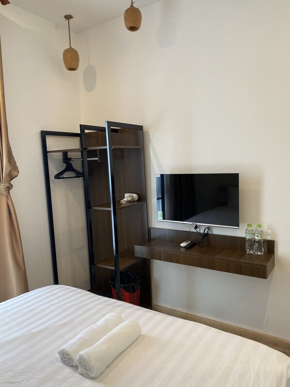 A hotel room with a bed, a TV, a shelf with bottles, a coat rack, and a trash bin.