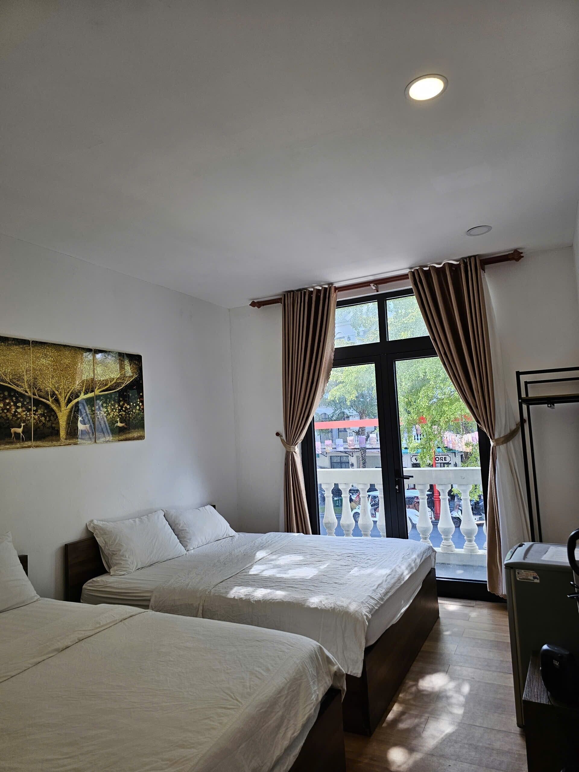 A hotel room with two beds, a ceiling light, a window with curtains, a balcony, and a painting on the wall.