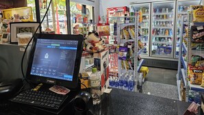 Una caja de pago con una computadora, un reloj digital y una variedad de aperitivos y bebidas al fondo.