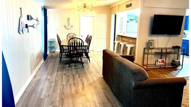 A living room with a wooden floor, a sofa, a dining table with chairs, a chandelier, and a TV mounted on the wall. - Blue Oasis (Marion)