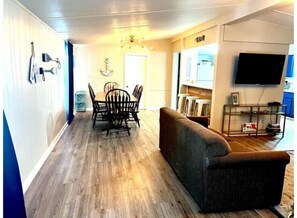 A living room with a wooden floor, a sofa, a dining table with chairs, a chandelier, and a TV mounted on the wall. - Blue Oasis (Marion)