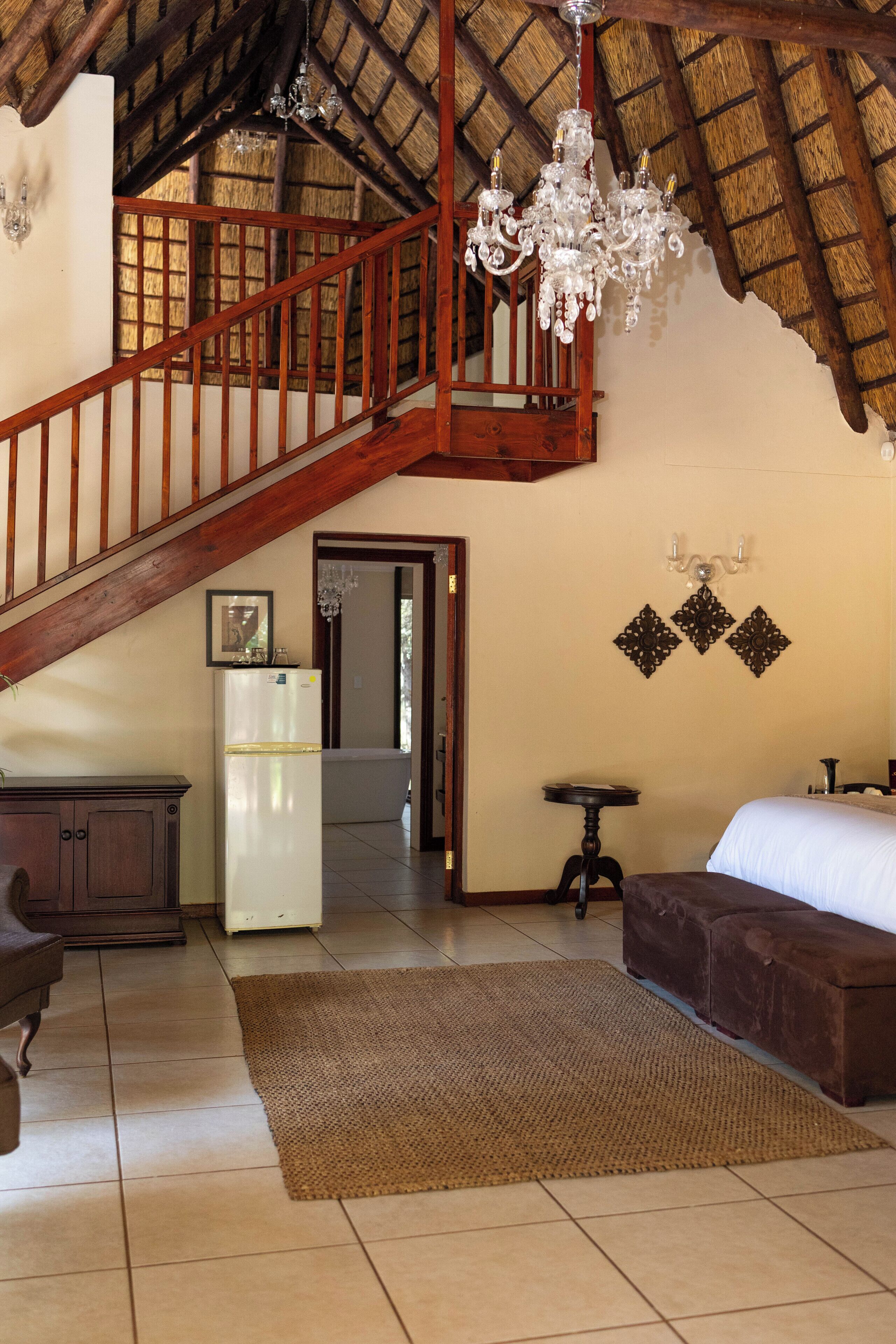 A spacious room with a high ceiling, a wooden staircase, a chandelier, a refrigerator, a bed, and a small table.