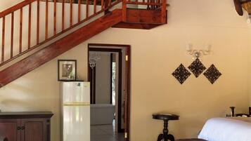 A spacious room with a high ceiling, a wooden staircase, a chandelier, a refrigerator, a bed, and a small table.