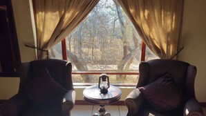 Two armchairs facing each other with a small table between them, a lantern on the table, and a view of trees through a window with curtains. - Phakela Private Lodge (Thabazimbi)