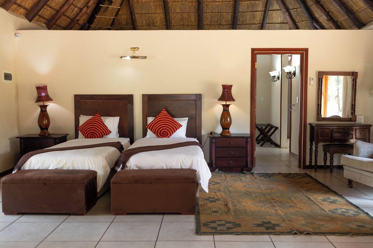 A room with two beds, a wooden ceiling, a mirror, a desk, and a patterned rug.