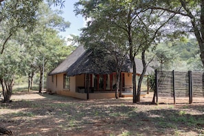 Un bâtiment rustique, doté d’un toit de chaume, entouré d’arbres et bordé d’un sol en terre battue.