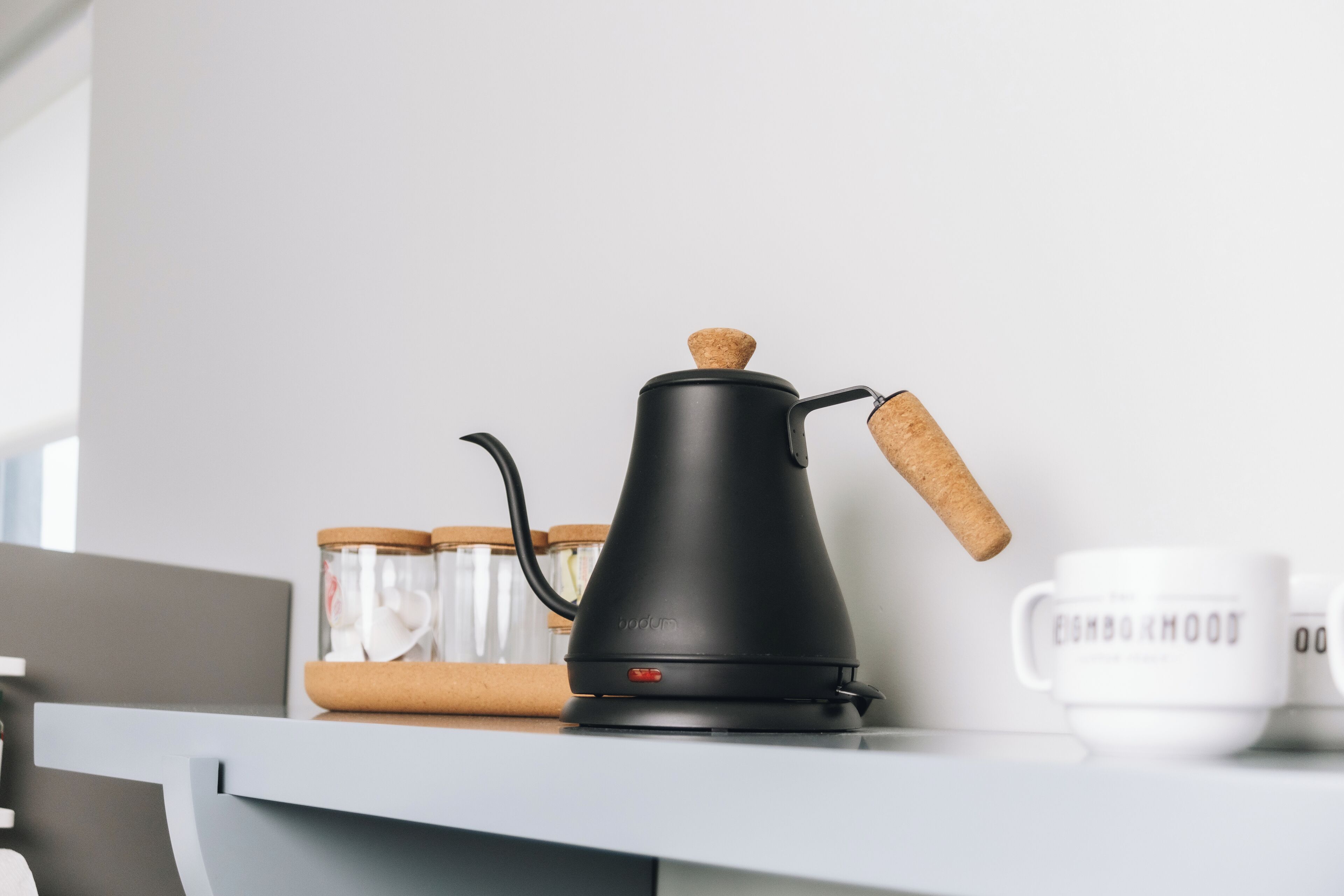 Ein schwarzer Wasserkocher mit Korkgriff auf einer weißen Arbeitsplatte, zusammen mit zwei Glasbehältern mit Korkdeckeln und einer weißen Tasse mit Text.