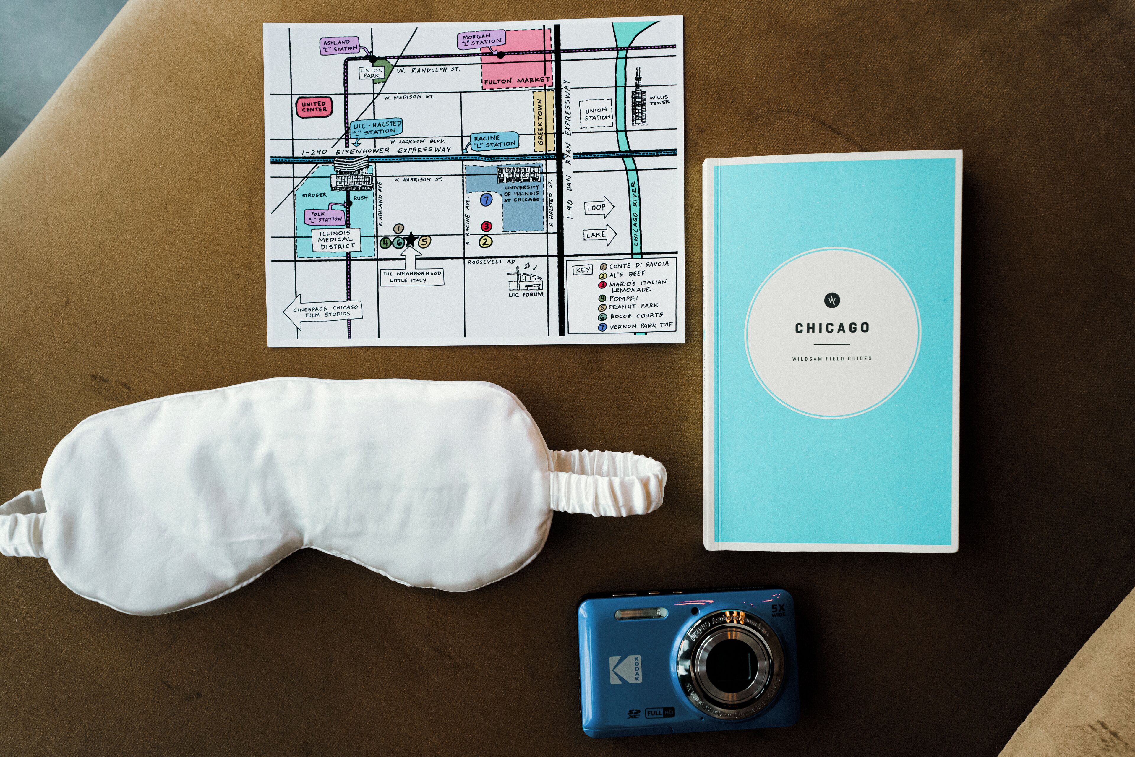 A map, a book titled 'CHICAGO,' a white pillow, and a blue camera on a brown surface.
