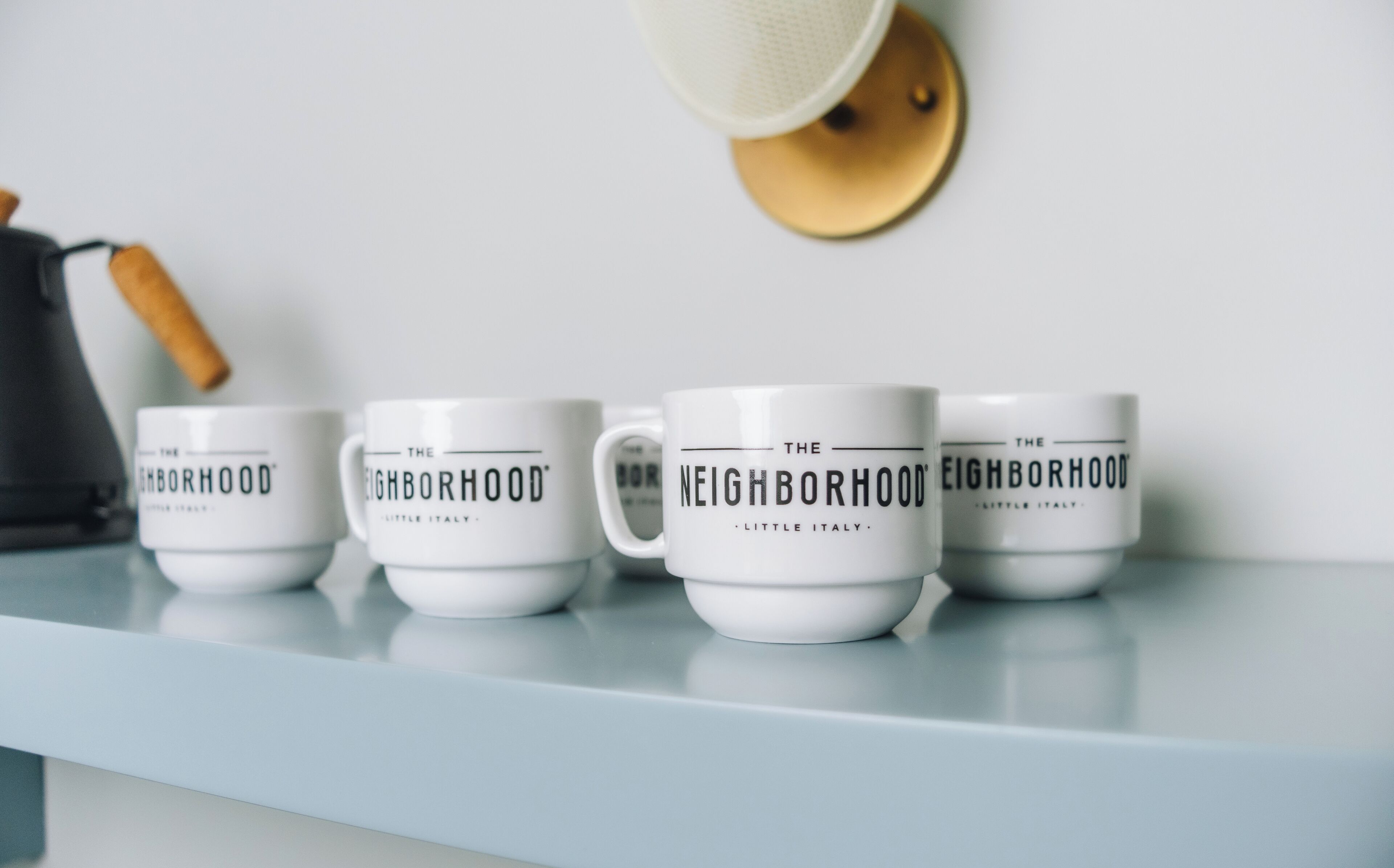 Four white mugs with "The Neighborhood" text on a light blue shelf.