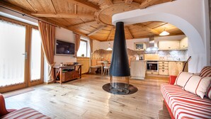 A cozy living room with a fireplace, wooden flooring, and a sofa. A kitchen with wooden cabinets and a dining area is visible in the background. - 183A Cristina Hospitality (Ortisei)