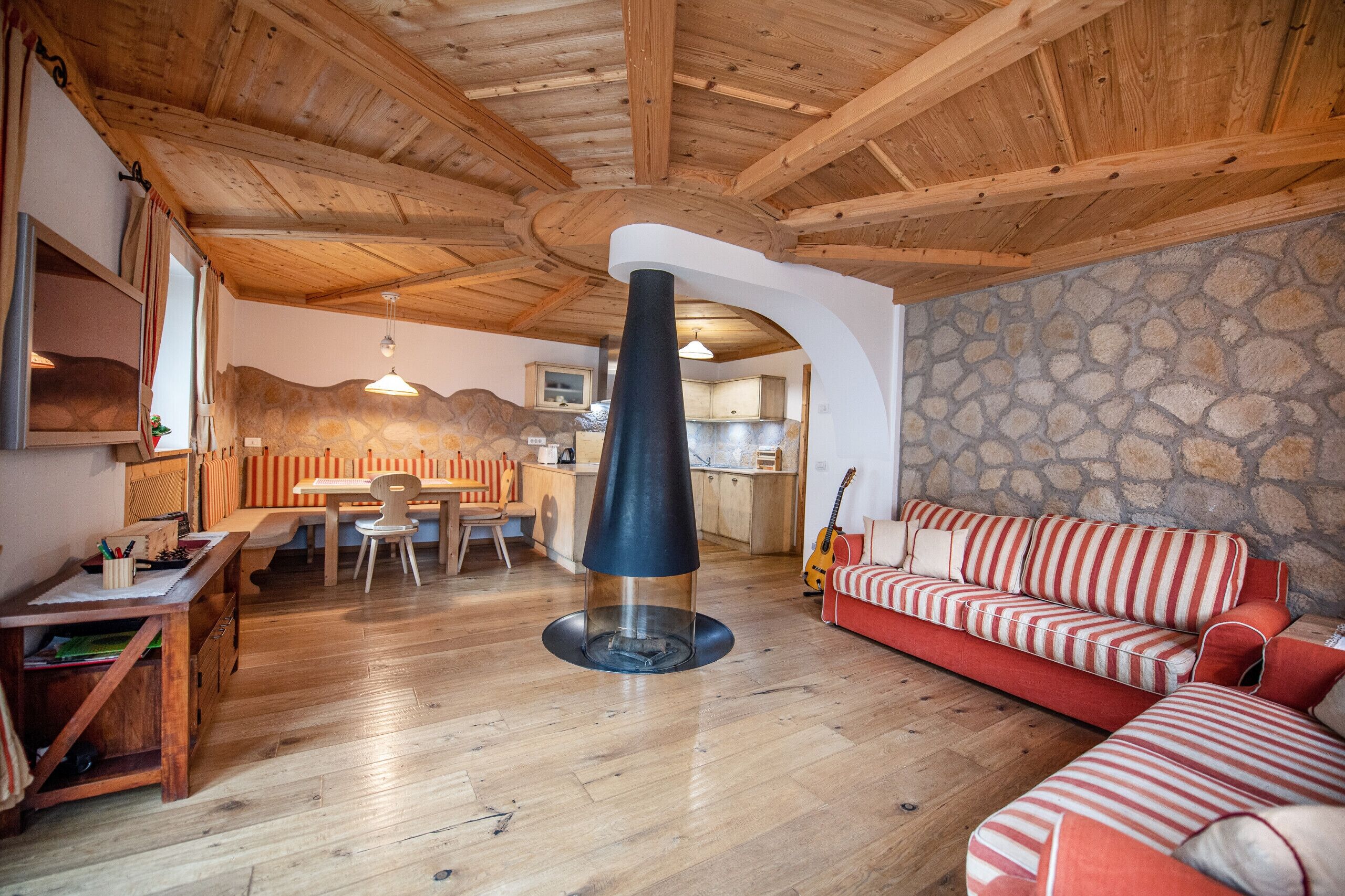 Ein gemütliches Wohnzimmer mit Steinwand, Holzbalkendecke, Kamin, einem rot gestreiften Sofa und einer Essecke mit Holztisch und Stühlen.