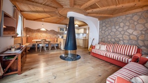 A cozy living room with a stone wall, wooden ceiling beams, a fireplace, a red-striped sofa, and a dining area with a wooden table and chairs. - 183A Cristina Hospitality (Ortisei)