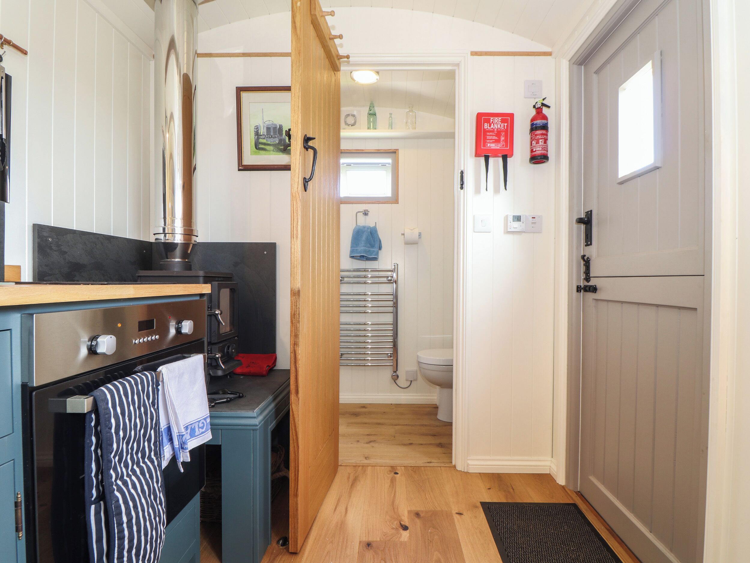 A compact kitchen with a stove, oven, and a wooden door leading to a bathroom with a toilet and a fire extinguisher.