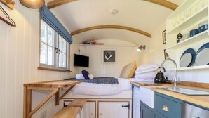 A compact kitchen with a bed, wooden stools, and a built-in bench. - Bosulla Shepherds Hut (Penzance)