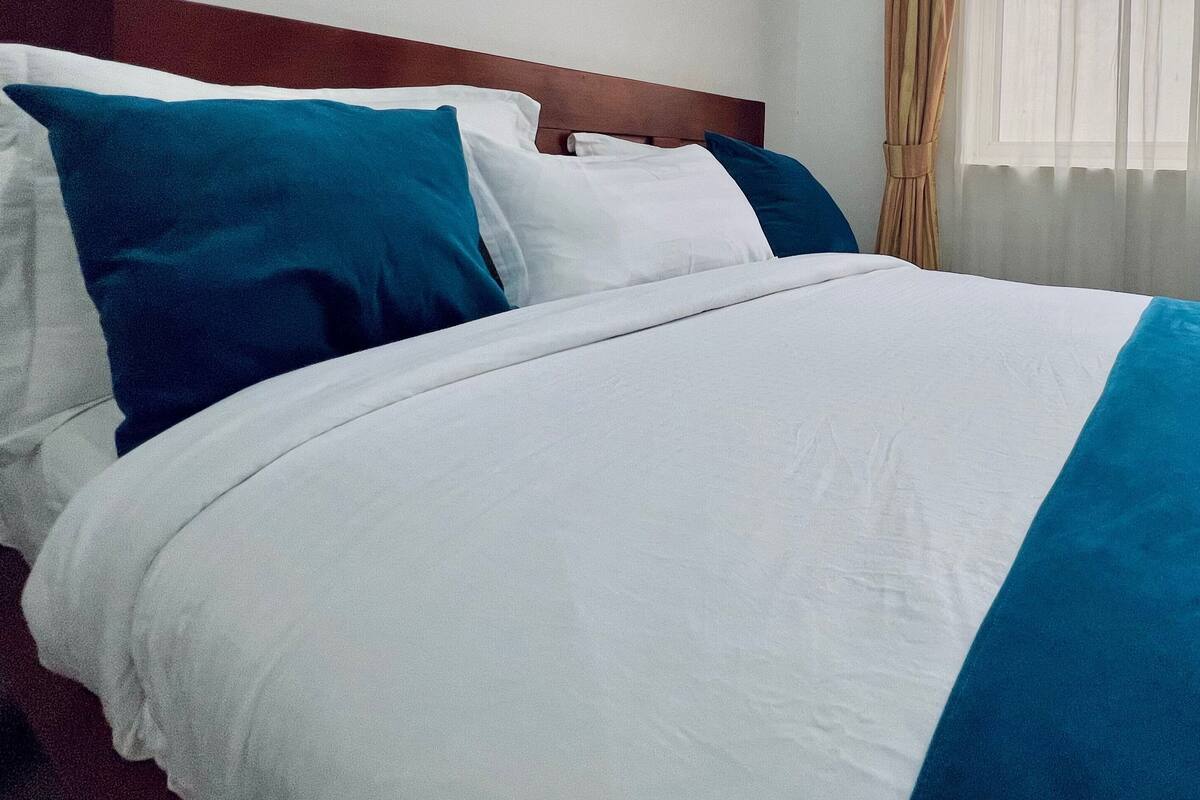 A neatly made bed with white and blue bedding, a wooden headboard, and a window with sheer curtains.