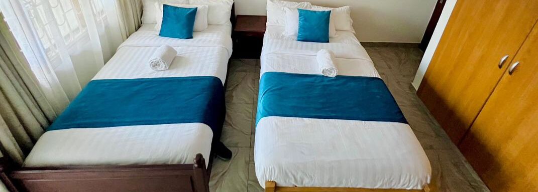 Two single beds with blue and white bedding, a wooden wardrobe, and a tiled floor.