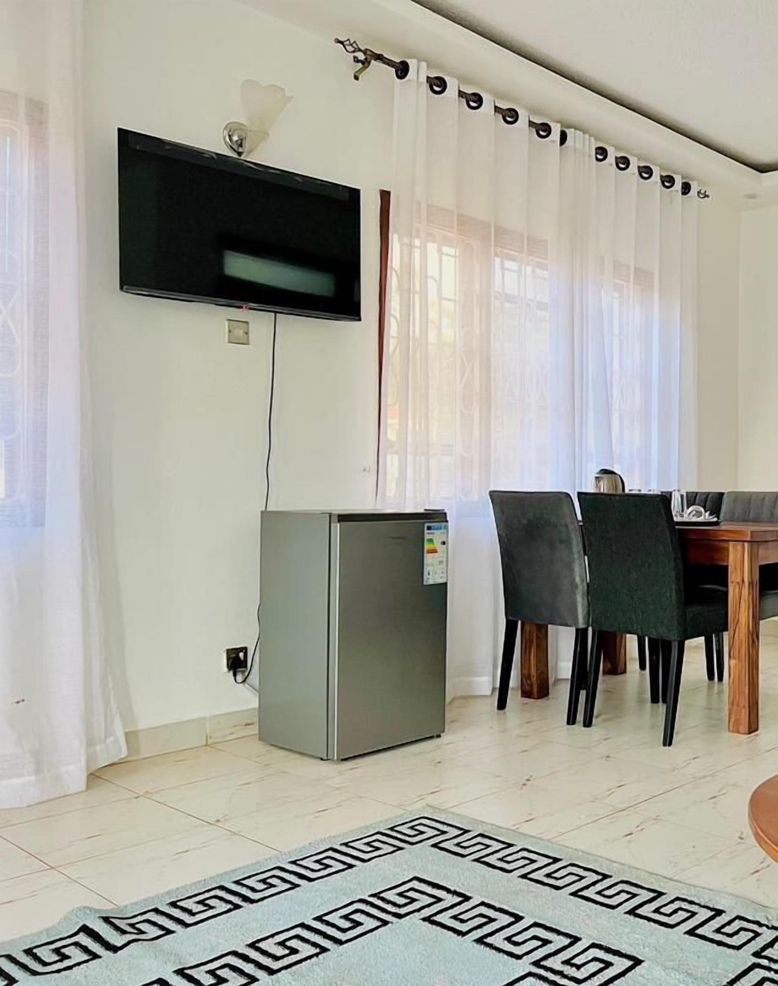 A living room with a flat-screen TV mounted on the wall, a refrigerator, a dining table with chairs, and a patterned rug.