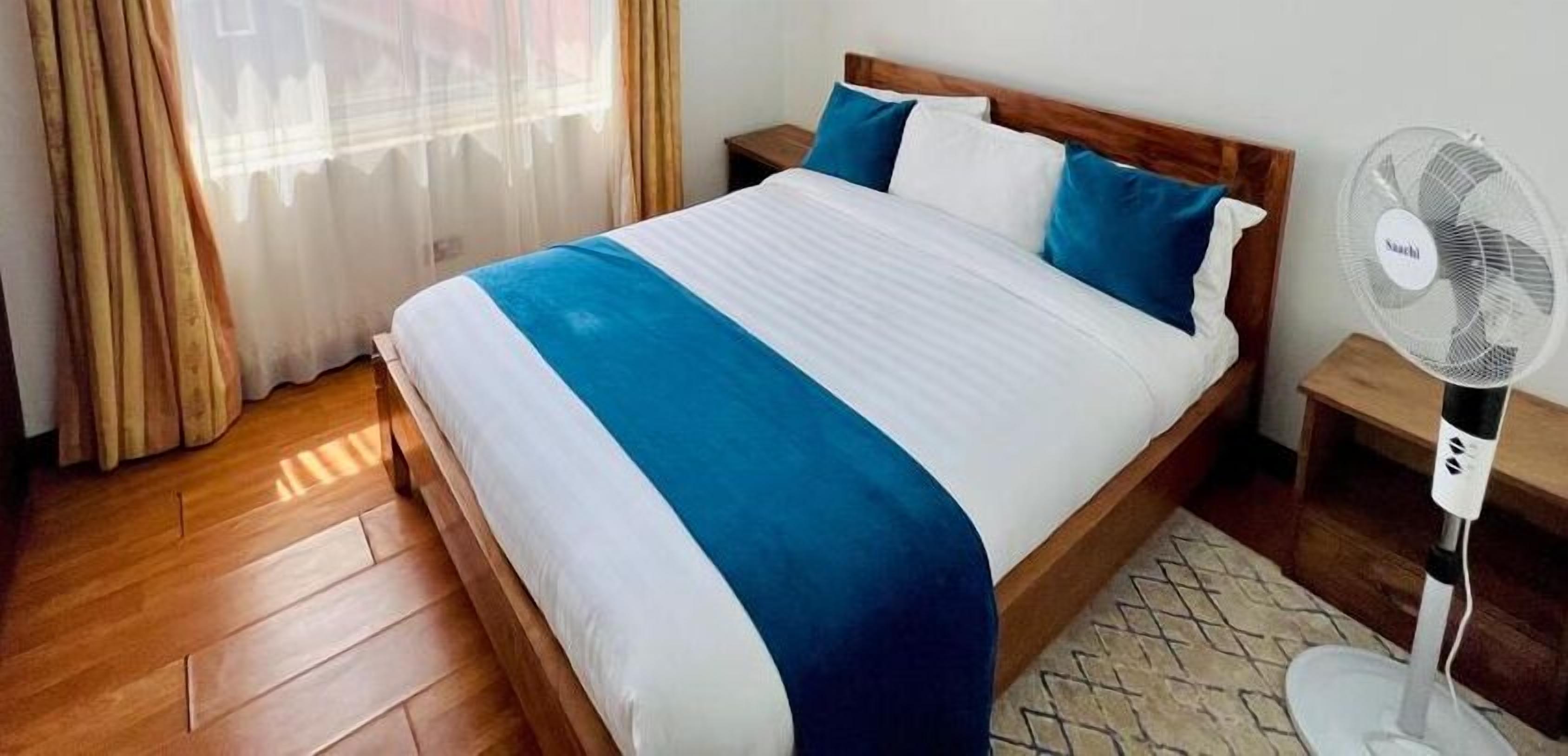 A neatly made bed with a blue and white bedspread, a wooden headboard, and a white fan on a wooden nightstand.
