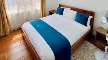 Una cama bien tendida con colcha azul y blanca, cabecera de madera y un ventilador blanco sobre una mesita de noche de madera.