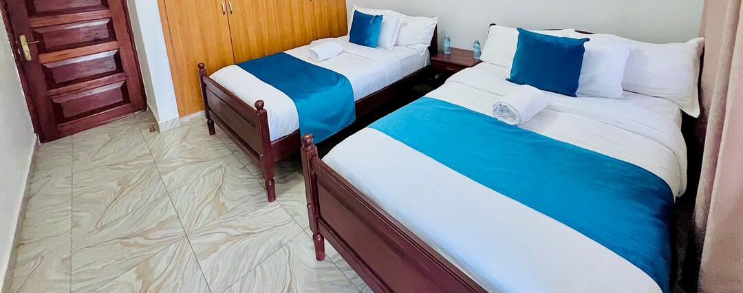 A hotel room with two single beds, each with a blue and white bedspread, wooden headboards, and a wooden wardrobe in the corner.