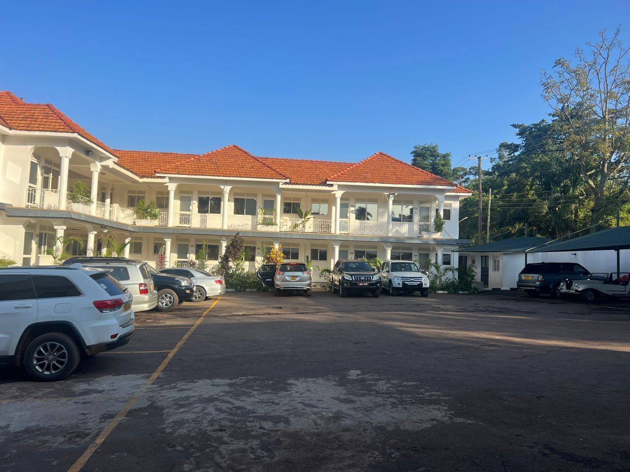 A multi-story hotel with a red-tiled roof, a parking lot with several cars, and a covered area on the right.