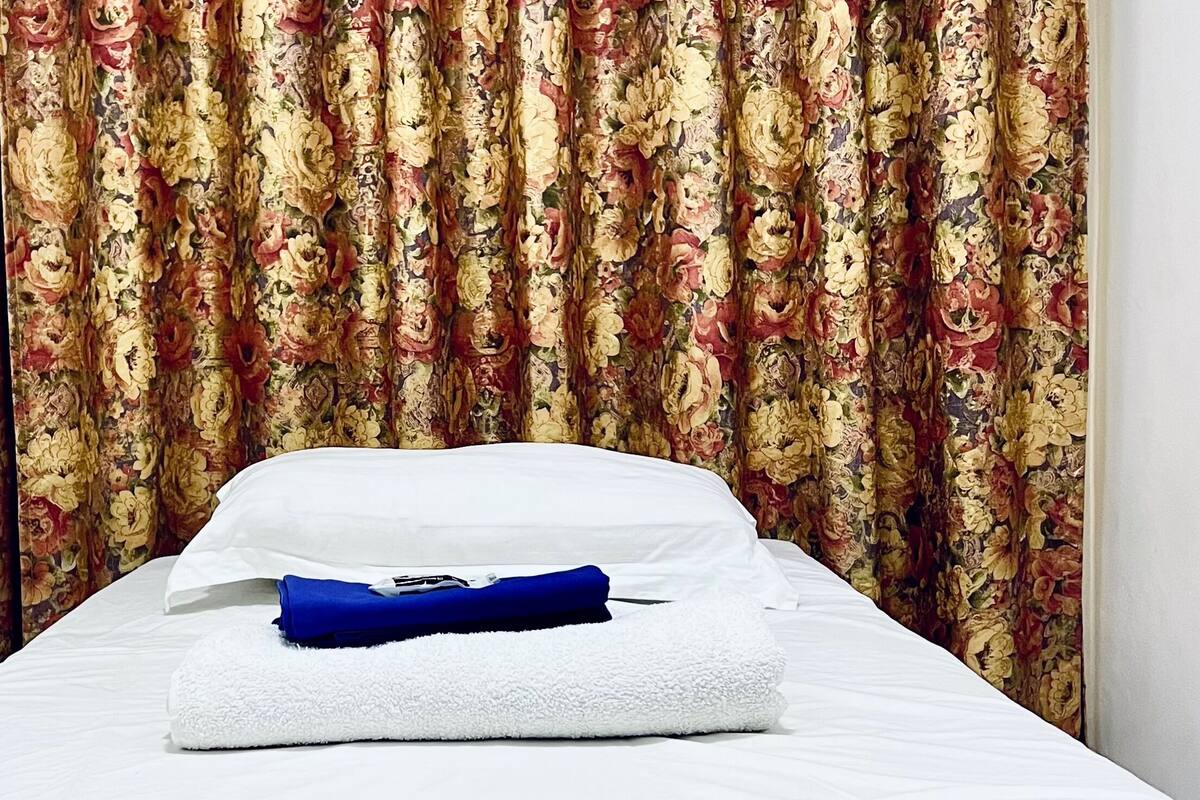 A neatly made bed with white sheets and a blue blanket, a folded blue towel, and a floral curtain in the background.