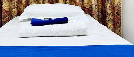 Una cama bien tendida con sábanas blancas y una manta azul, una toalla azul doblada y una cortina con estampado floral al fondo.