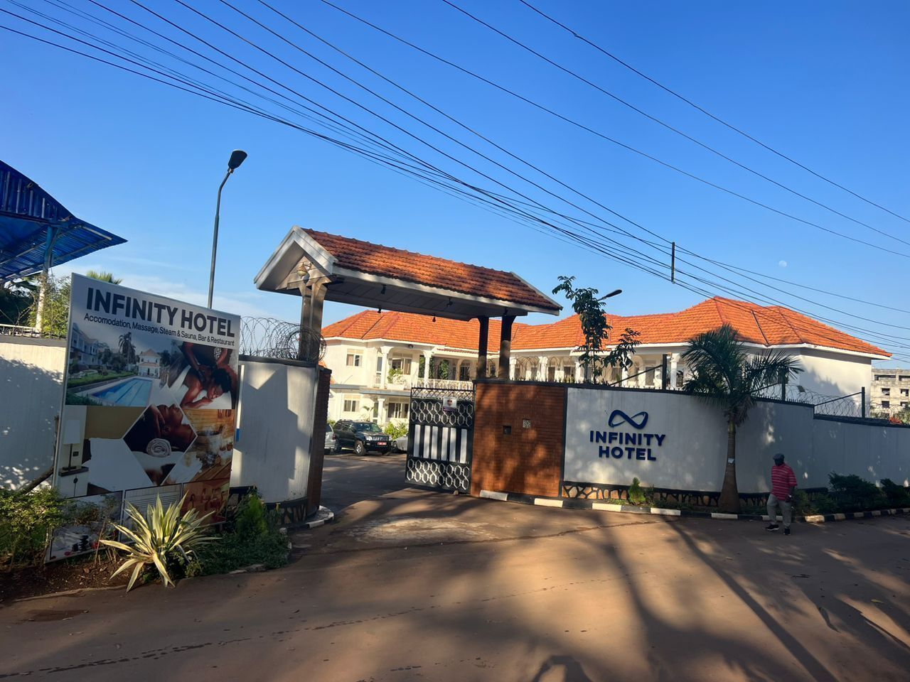 The Infinity Hotel entrance with a gated driveway, a large signboard, and a clear blue sky.