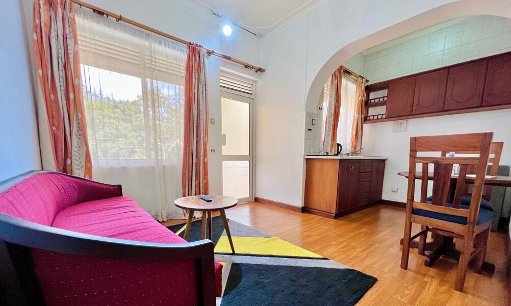 A cozy living room with a sofa, a small round table, and a wooden chair. There is a kitchen area with cabinets and a countertop.