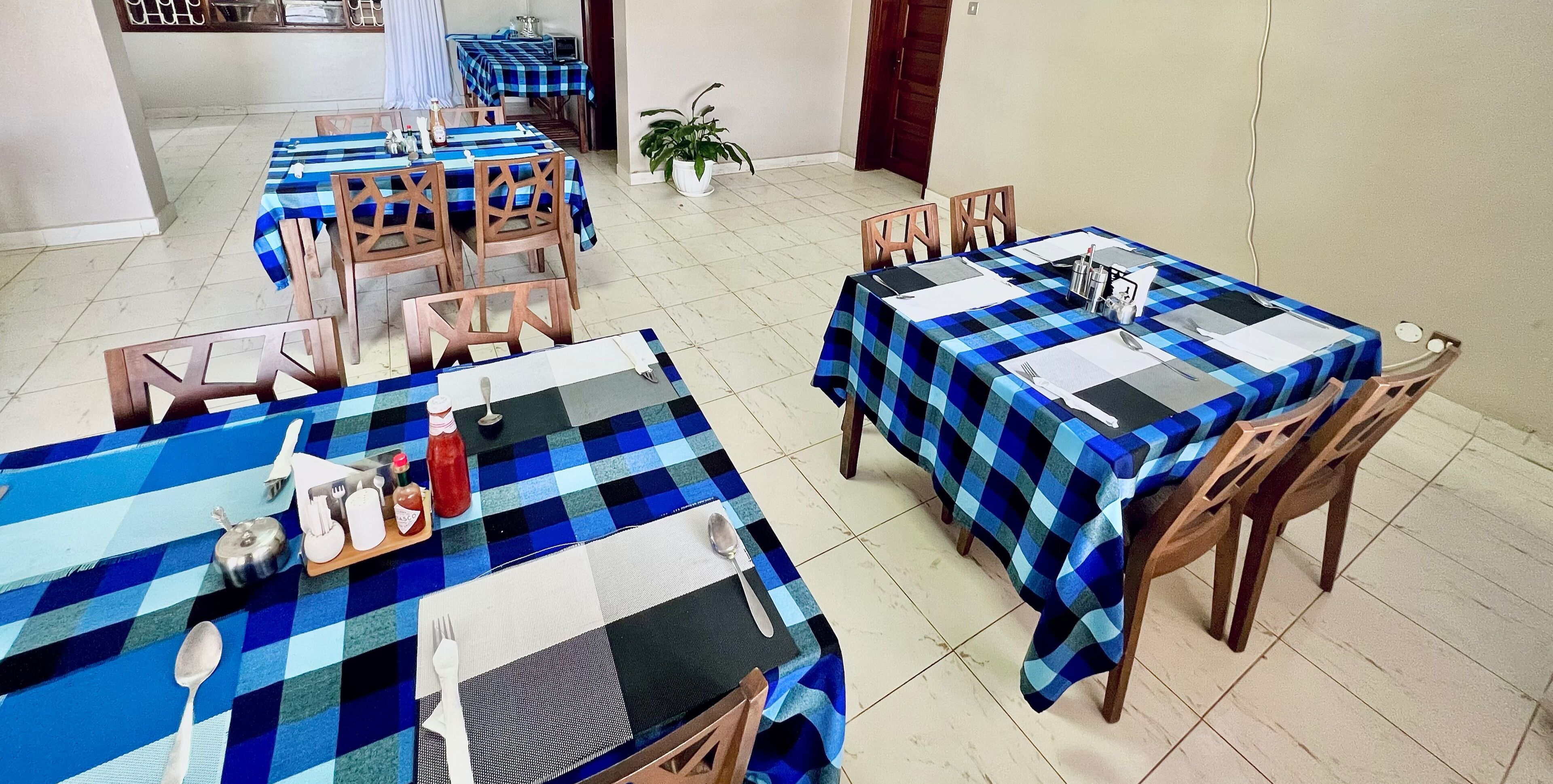 A dining area with multiple tables covered in blue and black checkered tablecloths, wooden chairs, and basic table settings including cutlery and condiments.